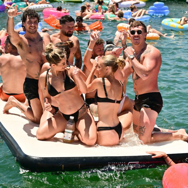 A group of people in swimsuits party on a floating platform in a lake, smiling and raising their hands. The water is filled with colorful inflatable tubes and other revelers. Boats from The Yacht Social Club Sydney Boat Hire are anchored in the background, adding to the lively atmosphere.