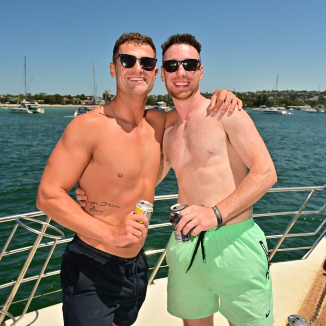 Two men in swimsuits and sunglasses smile at the camera while standing on a boat in sunny, open waters. One has his arm around the other, holding a drink. The background showcases other boats and a distant shoreline. Text on the image reads "Yacht Social," capturing their joyous moment at sea with friends.