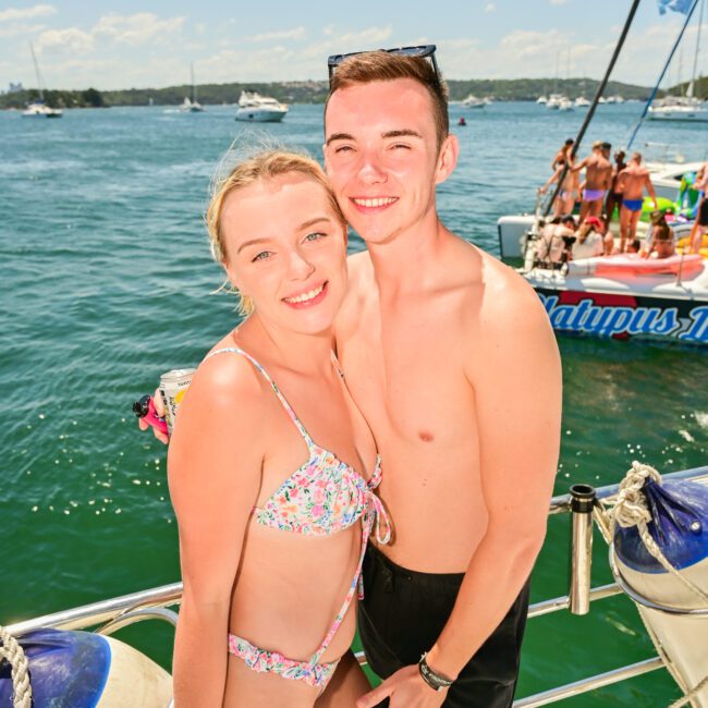 A young couple stands on a boat deck smiling at the camera. The woman is wearing a floral bikini, and the man is in black swim trunks, holding a phone to capture the moment. The background features a blue ocean with other boats and people on another vessel under the clear, sunny sky.