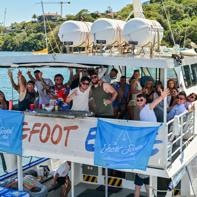 A group of people are gathered on the upper deck of a boat, enjoying a lively party. They are smiling, waving, and holding drinks. The boat is docked in a scenic location with lush greenery in the background. Two blue banners add to the festive atmosphere on the boat’s side rails.