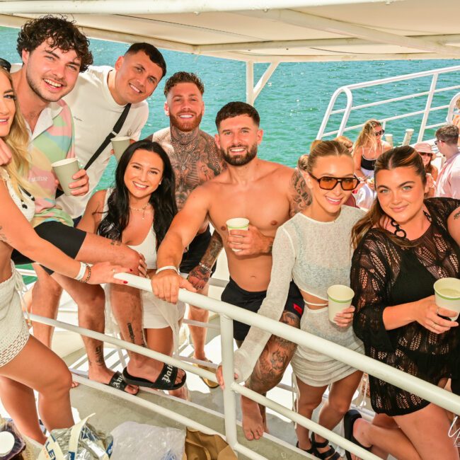 A group of nine people smiling and posing together on a boat. They are casually dressed in summer attire, some wearing swimsuits, and are holding drinks. The water and parts of the sailboat are visible in the background, suggesting a lively and enjoyable outing.