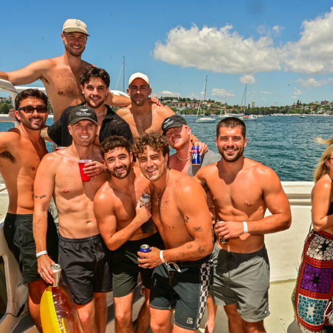 A group of shirtless men and some women are posing on a boat under a sunny sky. The men have drinks in hand and are smiling, with sailboats and Sydney Harbour's shoreline in the background. Some women are seen on the right side also enjoying The Yacht Social Club event.