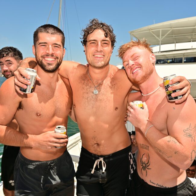 Three shirtless men are standing close together on a boat, smiling and holding cans. Two men are hugging the man in the center. Nearby, other people in swimwear can be seen enjoying the sunny day on the tranquil water.