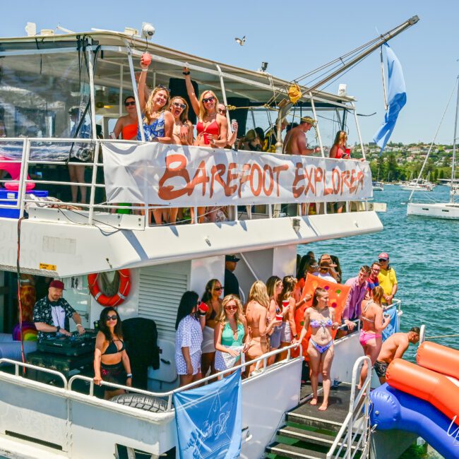 A lively party is taking place on a docked boat named "Barefoot Explorer." People are dancing, posing, and enjoying the sunny day. The boat is decorated with a festive banner, surrounded by inflatables in the water. Nearby boats and the scenic shoreline create a picturesque backdrop.