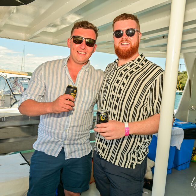 Two men on a boat smile while holding canned drinks. They are wearing sunglasses and casual short-sleeved shirts, one in stripes and the other in a patterned shirt. The serene background showcases a sunny day with blue skies and boats on the water.