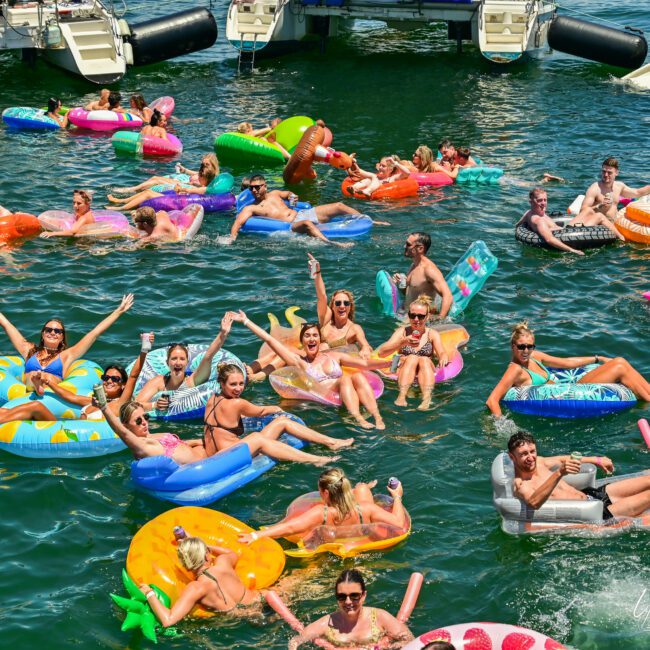 A group of people enjoying a sunny day while floating on various colorful inflatable rafts and tubes in a large body of water. Some are waving at the camera and smiling, with boats and yachts visible in the background. The atmosphere is festive and lively, perfect for making cherished memories.