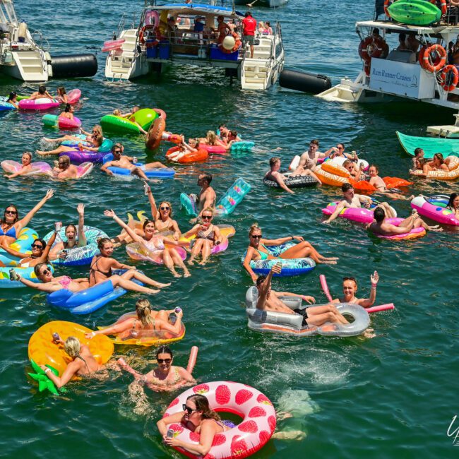 A group of people enjoying a sunny day on the lake with vibrant inflatables, including donuts, pineapples, and animals. They are swimming and relaxing near sleek yachts. The atmosphere is lively and festive with the "Yacht Social Club" logo visible in the background.