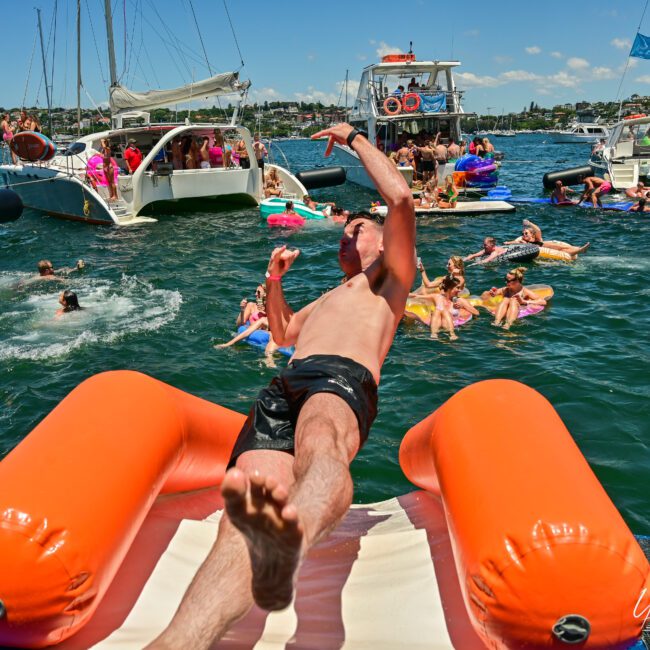 A man in swim trunks glides down an inflatable water slide off a boat into a lake, surrounded by numerous people swimming and floating on colorful inflatables. Several boats are gathered in the background with people enjoying the festive, sunny weather.