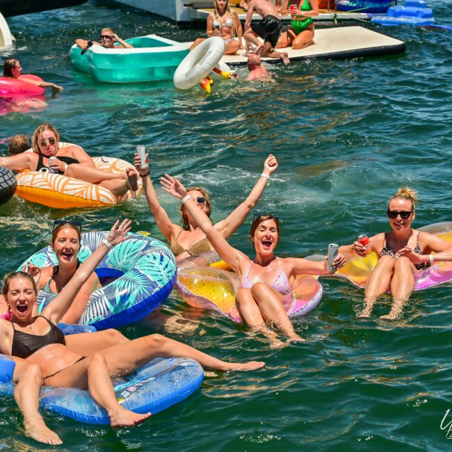 A group of people enjoying a sunny day on a lake, floating on various inflatable rafts. They are smiling, raising their arms, and holding drinks, creating a festive atmosphere. Other people can be seen in the background, also having fun on the water. "The Yacht Social Club Sydney Boat Hire" text on the image.