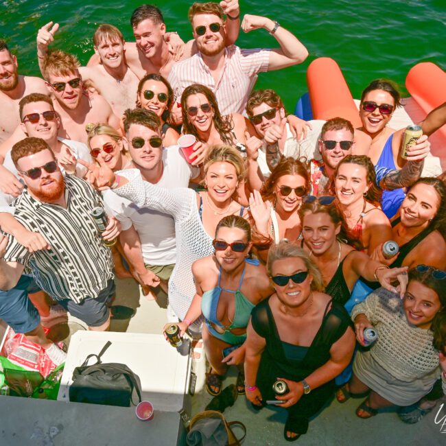 A large group of people in casual summer attire are smiling and posing for a photo on a boat with a green water background. Some are holding drinks and making playful gestures, while inflatable toys float nearby. A cooler with supplies is visible, enhancing the festive and jovial mood.