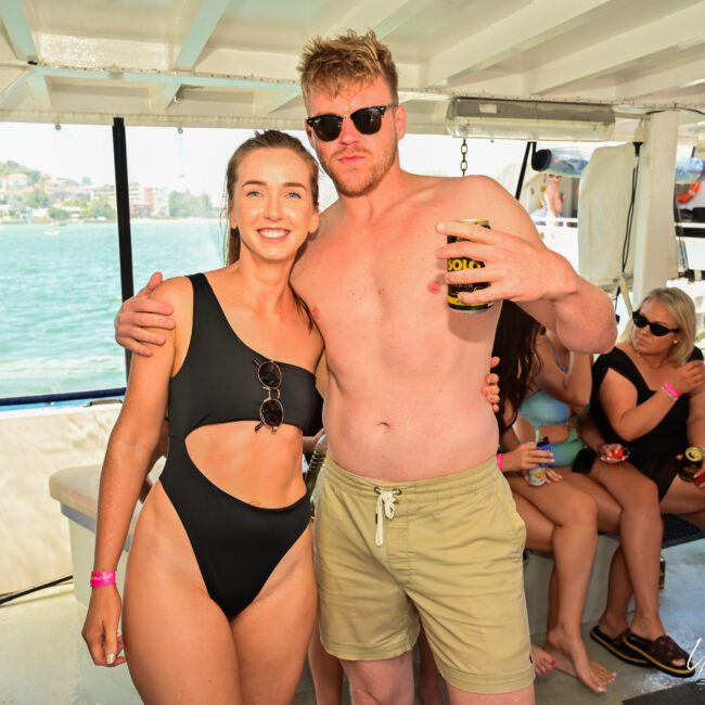 A man and woman, both in swimwear, stand arm-in-arm on a boat under the bright sun, each holding a beer. Water and land are visible in the background as other people clad in casual summer attire enjoy the lively boat ride.