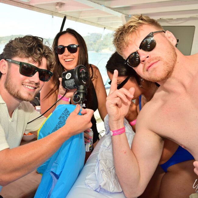 A lively group of four people are partying on a boat. Three shirtless men, wearing sunglasses, enjoy the moment while a woman holds a microphone and smiles. One man throws up a peace sign, adding to the joyous atmosphere as they have an unforgettable time on the water.