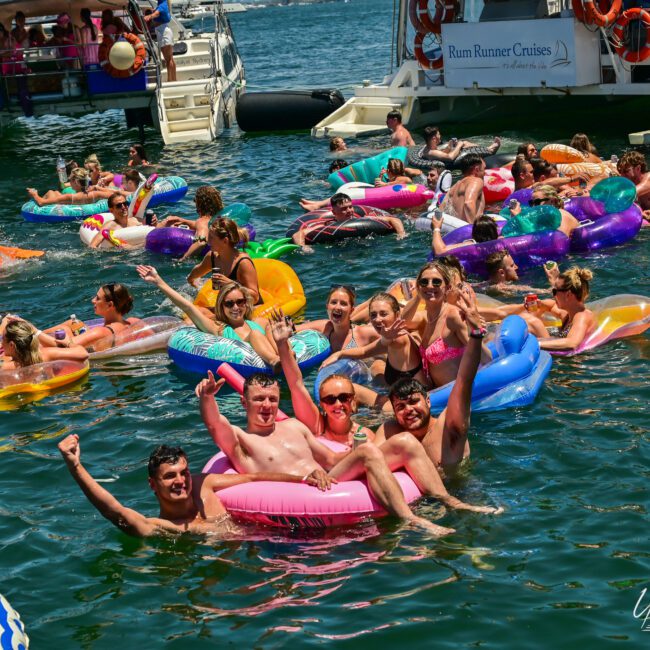 A large group of people enjoying a sunny day on the water, floating on inflatable rafts and inner tubes, with a festive atmosphere. They are smiling, raising their hands, and celebrating. Boats and pontoons anchor nearby, and there is a backdrop of other revelers and waterfront buildings.