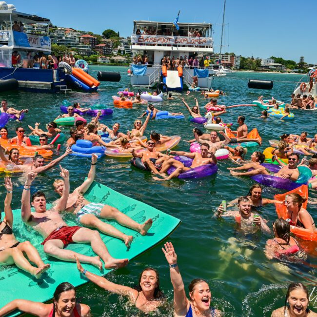 A lively crowd of people enjoying a sunny day at a large floating water party. They are swimming and floating on various inflatables and paddleboards in a body of water. Boats and yachts are docked nearby. The attendees are cheerful, waving, and socializing.