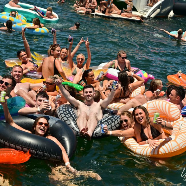 A large group of smiling people enjoying a sunny day and floating on colorful inflatable rafts in a lake. They are holding up drinks and cheering, creating a lively atmosphere. In the background, more people are seen savoring the festive water activity.