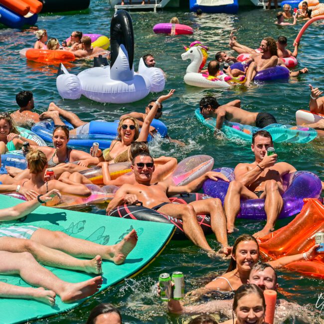 A lively group of people are having fun at a water party, floating on various inflatable rafts and toys, including a unicorn and swan. Many are sunbathing and enjoying tropical drinks, creating a festive atmosphere under the bright sun. A logo reads "Yacht Social.