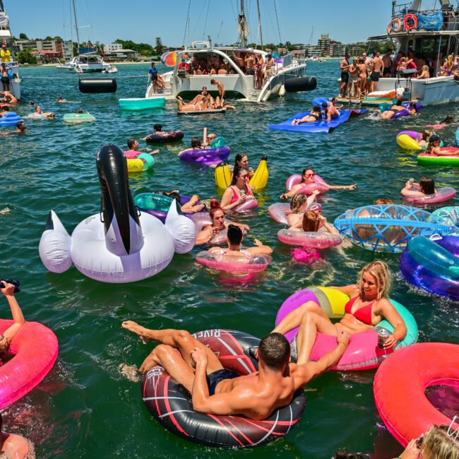 A lively scene of people enjoying a pool party on the water. They float on various colorful inflatable rings and rafts, including a large unicorn. In the background, boats are anchored, and the shoreline with buildings is visible under a sunny sky.