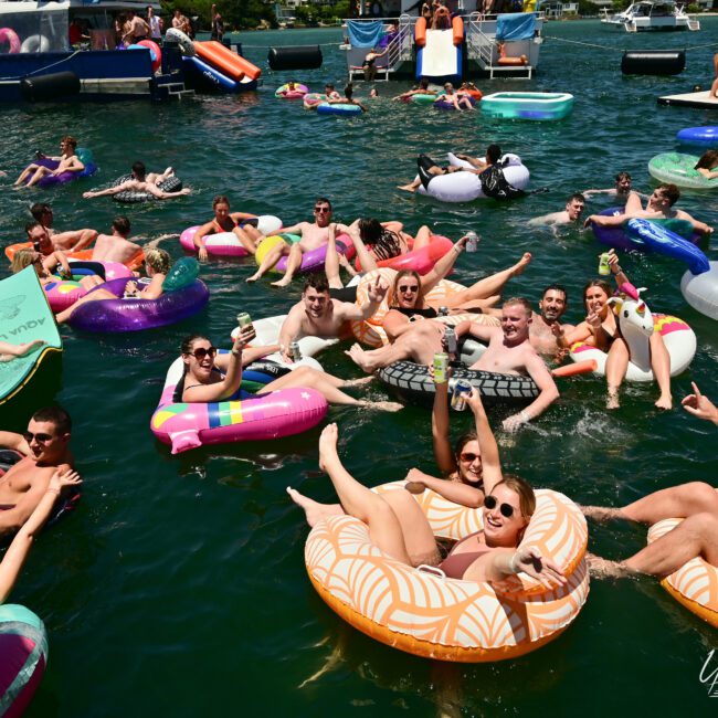 A crowded lake scene with numerous people enjoying themselves on colorful inflatable floats. The group is laughing, holding drinks, and socializing. Boats are visible in the background, and the atmosphere is lively and festive under a sunny sky, creating a vibrant summer escape by the lake.