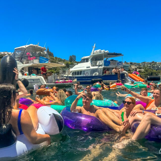 A lively scene at a boat party with numerous people relaxing on colorful inflatables in the water. Boats are docked in the background, and attendees are enjoying drinks and the sunny weather. The vibrant atmosphere is filled with socializing, laughter, and an overall festive vibe.