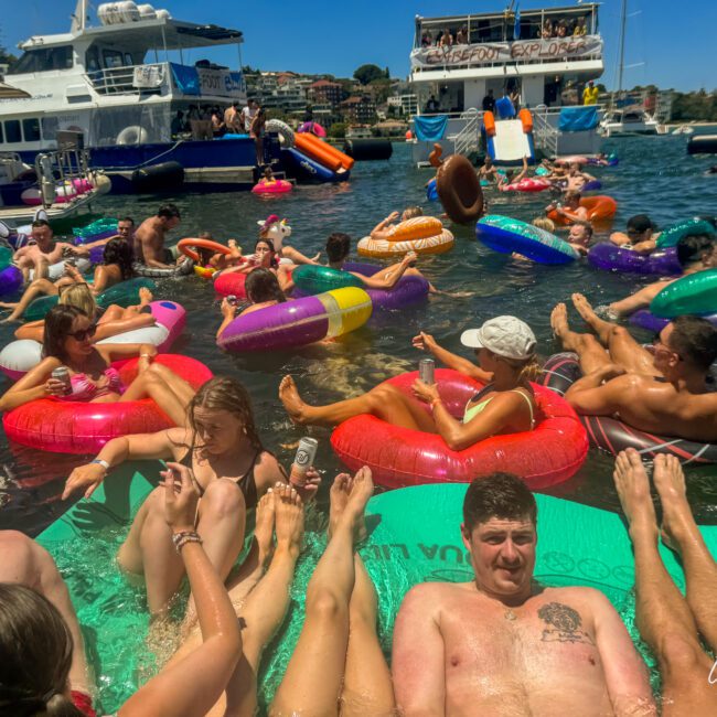 People are enjoying a sunny day on the water, floating in colorful kayaks and inflatable tubes. The background shows several boats and a dock with a crowd. The atmosphere is vibrant and festive, with people relaxing and socializing in the water.