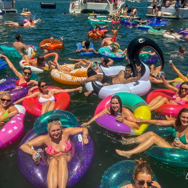 A large group of people enjoy a sunny day floating on inflatable pool toys in the water. They are smiling, relaxing, and holding drinks, with boats visible in the background. The colorful inflatables range from donuts to swans and other fun shapes like unicorns.