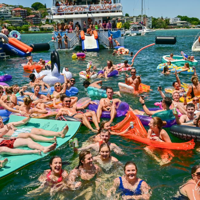 A large group of people enjoys a sunny day in the water, floating on various inflatable pool floats, including animals and simple mats. In the background, folks are aboard a vibrant boat named "Barefoot Explorer," diving into the fun. The atmosphere is lively and festive.