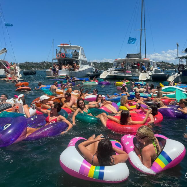 A large group of people enjoying a sunny day at a boat party, floating in the water on various colorful inflatable tubes. Several boats are anchored nearby, and the sky is clear and blue. The scene is vibrant and lively, with everyone having a fantastic time under the radiant sun.