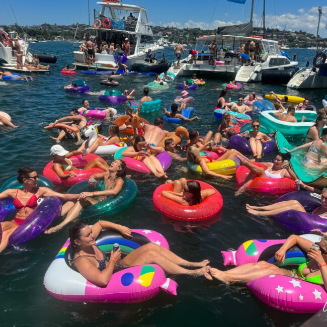 A lively group of people enjoying a sunny day on the water, each floating on colorful inflatable tubes. They are surrounded by several boats with more people on board. The scene is vibrant and festive, with smiles, drinks in hand, and upbeat music playing against a backdrop of a bright sky.