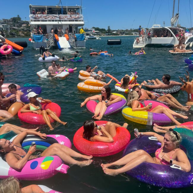 People are relaxing and having fun on colorful inflatable tubes and kayaks in the water by boats. A large party boat and a few sailboats are anchored nearby. Participants appear to be enjoying sunny weather and socializing.