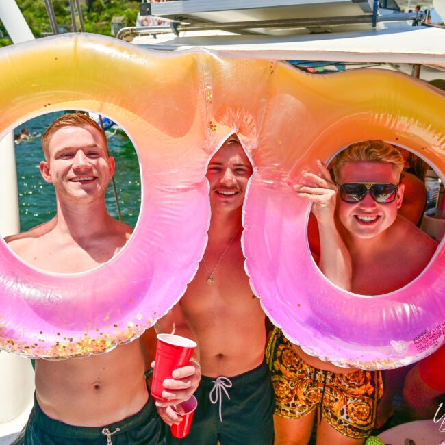 Three shirtless people posing with a large inflatable float on a boat. They are smiling and each holding red plastic cups. The background shows water and another boat enjoying the day. The multicolored float, in vibrant shades of pink and orange, adds to the lively atmosphere on this bright, sunny day.