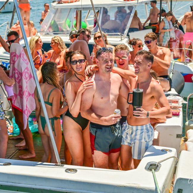 A group of friends in vibrant swimsuits are gathered on a boat, smiling and posing for a photo. Some hold drinks, while others chat and relax. The boat is lively with people enjoying the sunny day, with a few other boats visible in the background on the sparkling water.