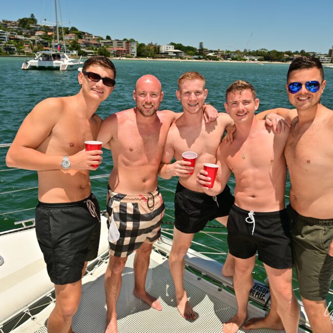 Five men are on a yacht in swimwear, holding red cups and smiling at the camera under a sunny sky. The backdrop features a serene body of water and a picturesque shoreline with houses, suggesting they are enjoying a pleasant social outing.