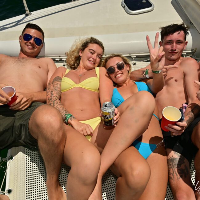 Five people are lounging on the deck of a boat, basking in the sunshine. Dressed in swimsuits, they hold drinks and smile widely. One person even makes a peace sign with their fingers, clearly savoring the joyful moment on the sparkling water.