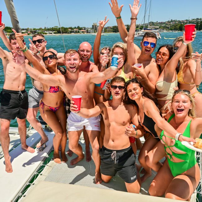 A group of people in swimwear are gathered on a boat, smiling and holding red cups. They appear to be having a fun time under the sunny weather near a picturesque waterfront with buildings and greenery in the background. Some have their arms raised in celebration, enjoying the perfect day on the water.