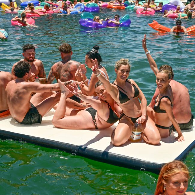 A group of people in swimsuits relax on a floating platform on a lake, smiling and waving at the camera. The water is filled with others enjoying colorful inflatables. A small logo in the corner reads "Desert Social.