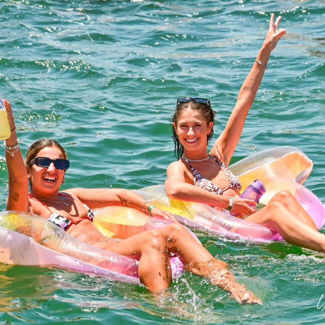 Two women are floating on inflatable pool loungers in a body of water, smiling and holding drinks. One woman wears sunglasses and holds a bottle, the other raises a peace sign. Both wear swimsuits as they enjoy the serene lake, with the water around them glistening in the sunlight.