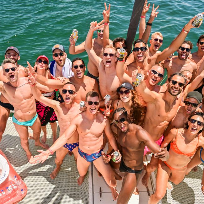 A joyful group in swimwear gathers on a boat, celebrating with drinks in hand. The background reveals open water, and a pink inflatable shaped like a rosé wine bottle bobs near the edge. Everyone's arms are raised in excitement.