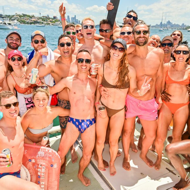 A large group of people in swimsuits joyfully pose for a photo on a boat under sunny skies. They are smiling, laughing, and holding drinks, with a backdrop of water and distant buildings. The atmosphere is lively and fun, perfect for a Luxury Yacht Rental Sydney experience.