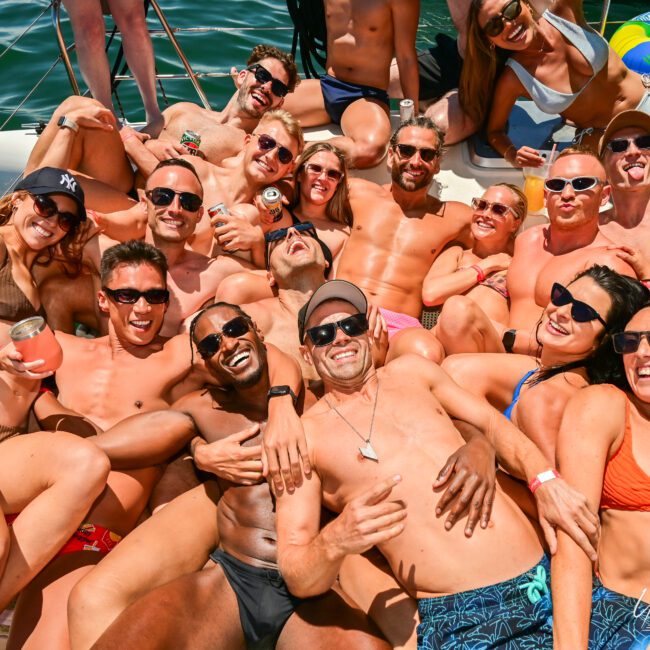 A group of people in swimsuits joyfully huddle together on a boat under sunny weather. Many are wearing sunglasses, smiling, and holding drinks. The boat cruises on the water, with various inflatable pool toys adding splashes of fun to the background.