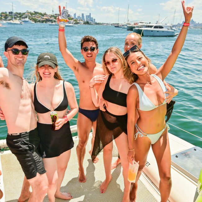 A group of six people in swimsuits are standing on a sailboat, smiling and posing for a photo. They are overlooking a body of water with several boats in the background. It's a sunny day with clear skies, and everyone appears to be enjoying themselves.