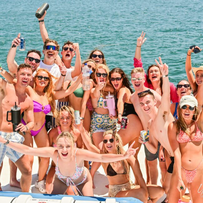 A group of about sixteen people, wearing swimsuits, are joyfully posing on a boat on a sunny day. They are smiling, raising their arms, and holding drinks, capturing the fun and excitement of a party or celebration on the water.