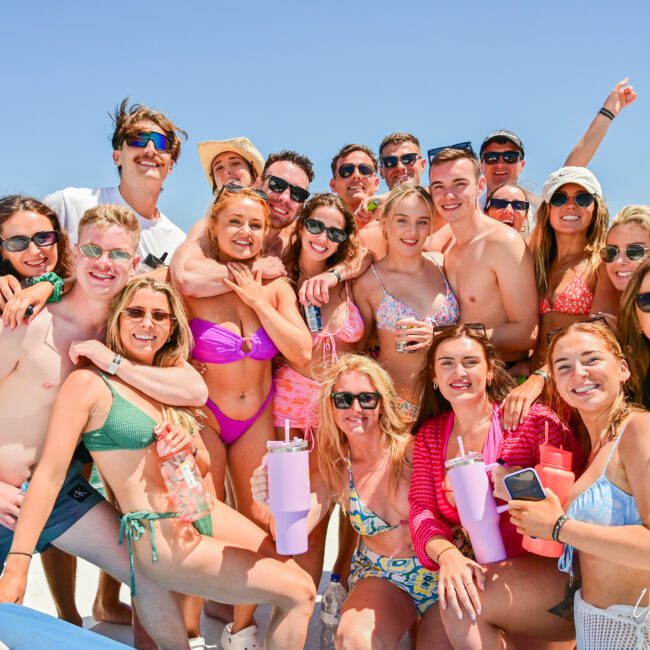 A group of cheerful people in swimsuits and summer attire gather closely together on a boat, posing happily for a photo. The sunny sky and calm water in the background suggest a day of fun and relaxation. Some hold beverages while smiling or laughing, sharing stories under the bright sun.