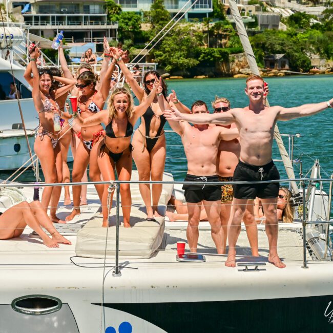 A group of people in colorful swimwear are joyfully posing on the deck of a sailboat. Some have their arms raised in celebration, while others are lounging and enjoying the sun. The background features sparkling water, lush greenery, and picturesque buildings. The atmosphere is lively and festive.