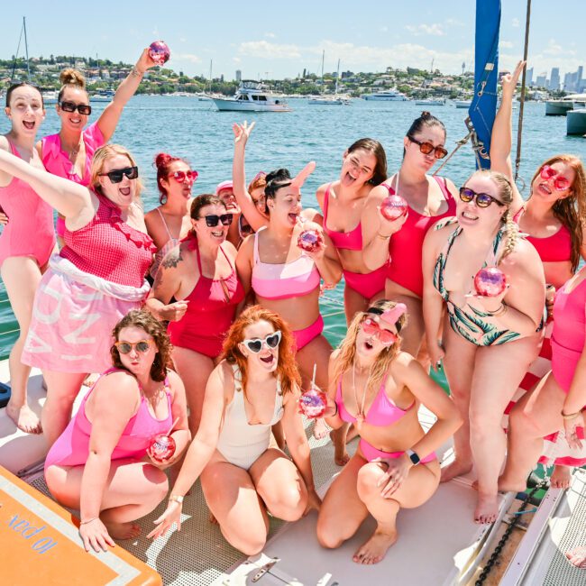 A group of people in bright swimwear posing cheerfully on a yacht under a sunny sky. Most are holding small pink beach balls, and the backdrop features a lively marina with several boats and a bustling city skyline. The atmosphere appears festive and joyful.