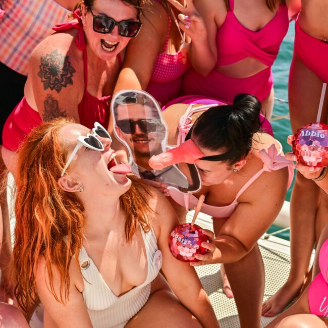 A group of women in matching pink swimsuits are celebrating on a boat. One woman holds a drink with "Abbie" written on it, while another playfully sticks out her tongue. The women are smiling, laughing, and enjoying themselves under the sun, with the clear blue sea in the background.