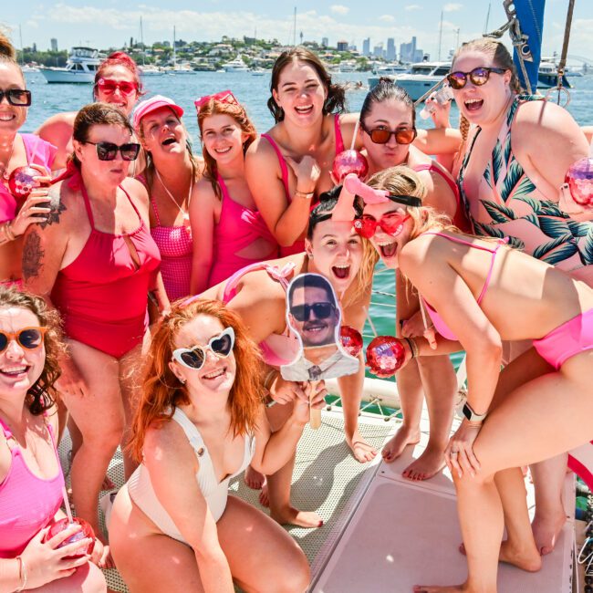 A large group of women dressed in red and pink swimsuits and accessories gather on the deck of a boat. They are smiling and posing for the camera on a sunny day. The cheerful scene unfolds against a backdrop of water, other boats, and a distant city skyline, capturing joyful moments under clear skies.