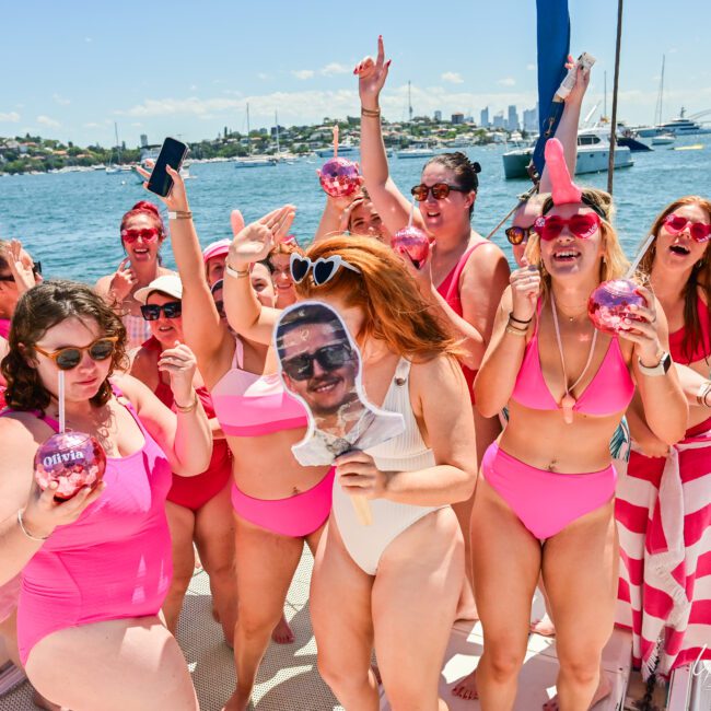 A group of people in pink swimsuits and dresses enjoy a boat party on a sunny day, with some holding drinks and one person holding a cutout of a face. They are celebrating with The Yacht Social Club Sydney Boat Hire against the backdrop of a harbor and cityscape in the distance.