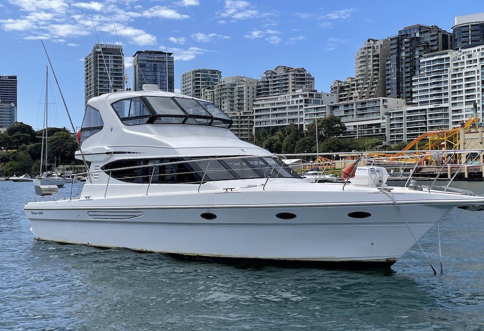 A sleek, white motor yacht is docked in a calm harbor. Tall buildings and a construction site with cranes form an urban backdrop. The sky is clear with scattered clouds, and the water beautifully reflects the scene.