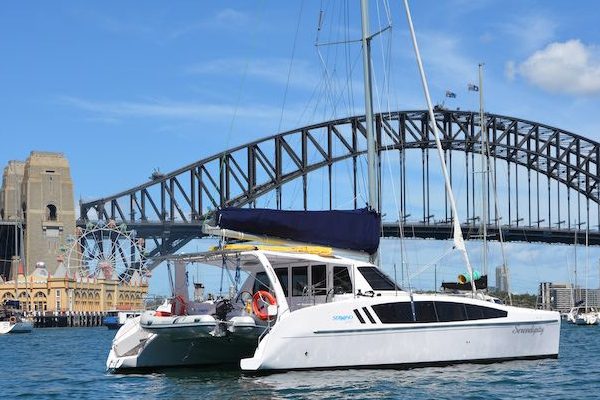 A catamaran named "Dreamtime" is anchored on calm water with a bridge arching in the background under a blue sky. The bridge features a large Ferris wheel on one side, hinting at an amusement park nearby. Several serene sailboats are also visible in the scene, adding to the picturesque setting.