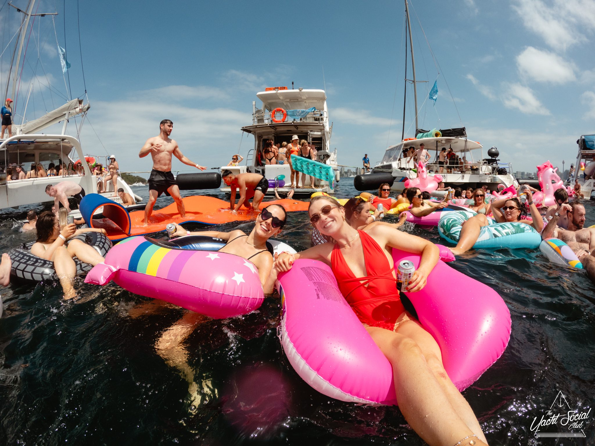 A lively group enjoys a day on the water, floating on colorful inflatable rafts near several boats. They're relaxing, laughing, and socializing under a bright, clear sky. Some hold drinks while others stand or swim in the water—an ideal scene from The Yacht Social Club's luxury yacht rentals in Sydney.